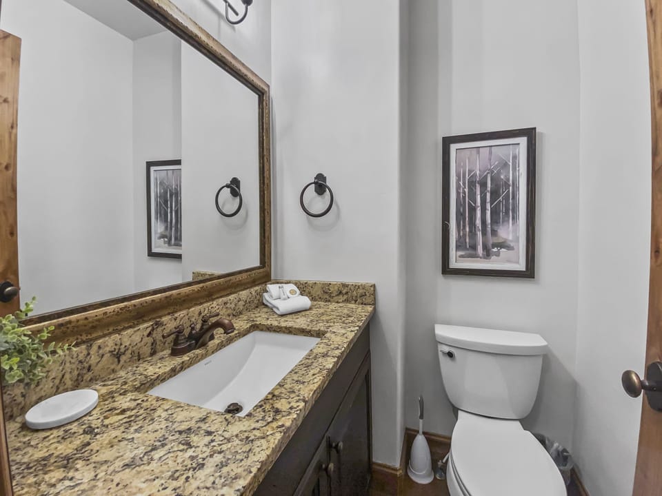 This elegant powder room features a beautiful granite vanity, dark wood cabinetry, and a large wood-framed mirror. Charming nature-themed artwork and soft lighting create a refined, welcoming space.