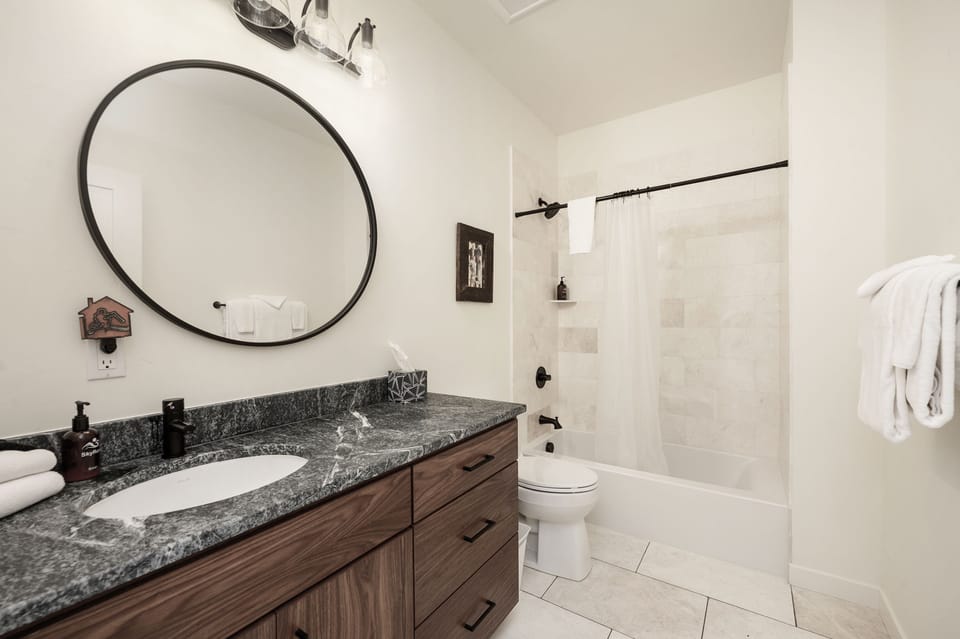 - Modern bathroom with granite countertop and stylish wood cabinetry   - Round mirror and bright lighting create a clean and fresh atmosphere   - Spacious shower and tub combo provide comfort after outdoor activities