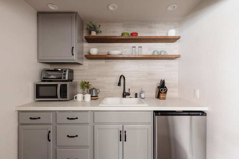 A modern kitchen with gray cabinets, a white countertop, a sink, and wooden shelves. It includes a microwave, toaster oven, small dishwasher, and a knife block.