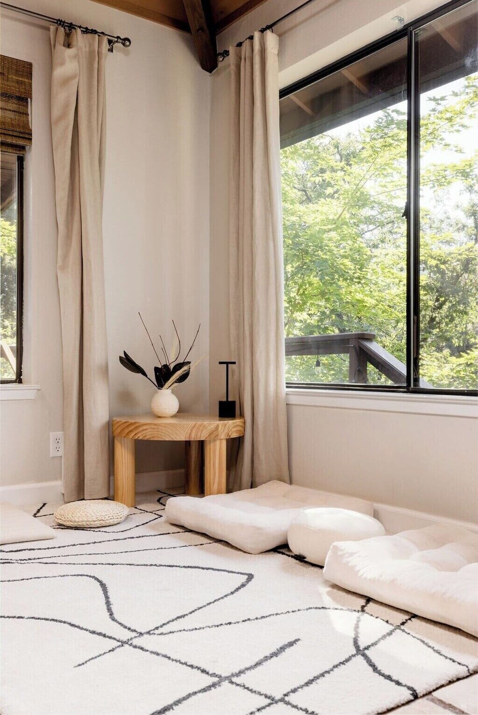 A cozy corner with a small wooden table, floor cushions, and a patterned rug by a window with beige curtains. A vase with decorative branches is on the table.
