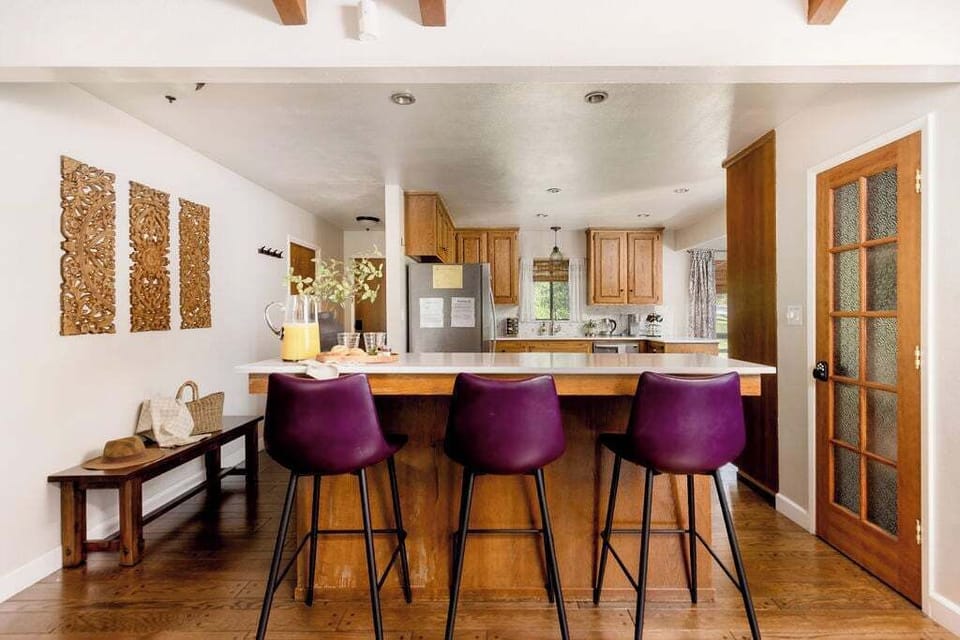 A bright kitchen with a central island, three purple bar stools, wooden cabinets, and stainless steel appliances. Decorative wooden panels and a bench are on the adjacent wall.