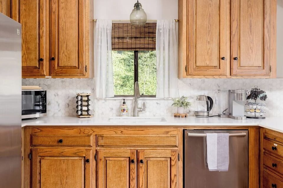 A kitchen with wooden cabinets, a white countertop, and a stainless steel sink. A window with bamboo shades and white curtains is above the sink. Appliances include a microwave, kettle, coffee maker, and dishwasher.