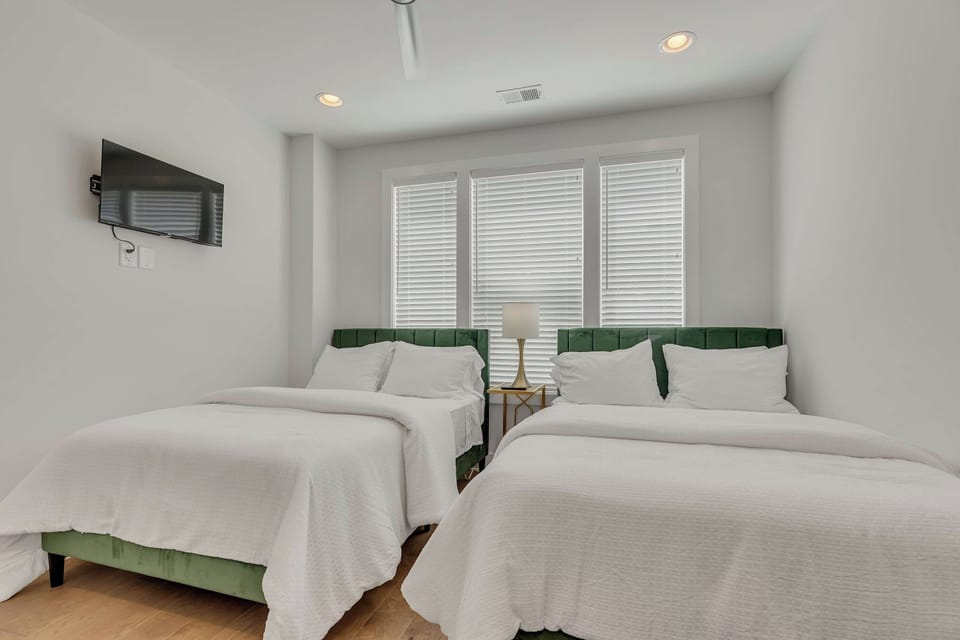 Bright bedroom featuring two plush beds with green upholstered headboards, crisp white linens, and large windows with natural lighting.