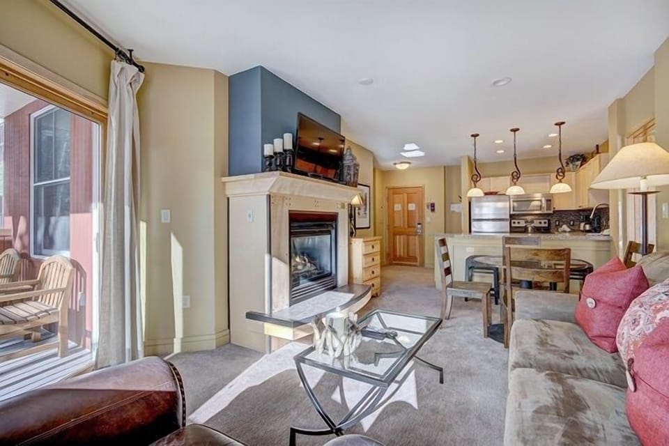 A cozy living room with a fireplace, glass coffee table, and beige sofa, adjacent to a kitchen area with a breakfast bar and stainless steel appliances. A sliding door opens to a patio with chairs.