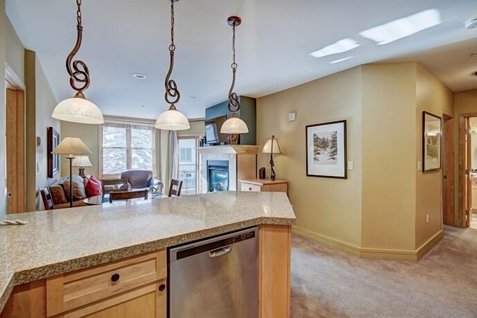A modern kitchen with granite countertops, stainless steel dishwasher, and pendant lights opens to a cozy living area featuring a fireplace, armchairs, and large windows with a scenic view.