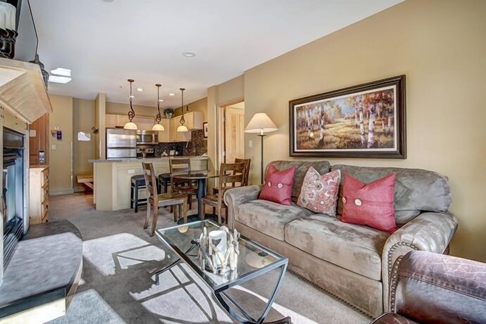 A cozy living room with a brown sofa, red cushions, a glass coffee table, and a wall painting. The setup includes a connected kitchen area with bar stools and pendant lighting.