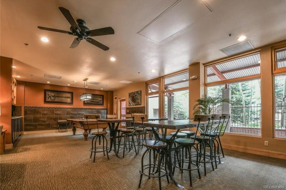 A spacious dining area with wooden tables and chairs, ceiling fans, and large windows providing natural light. Decor includes wall art and a potted plant.