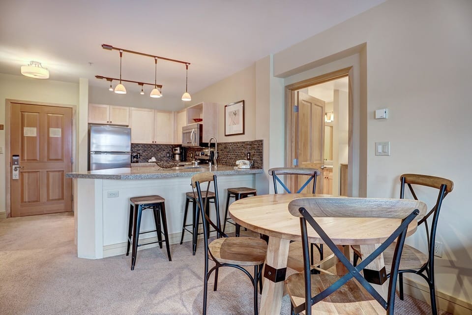 A modern kitchen with light wooden cabinets, stainless steel appliances, and an island with stools, adjacent to a round dining table with four chairs in a well-lit room.