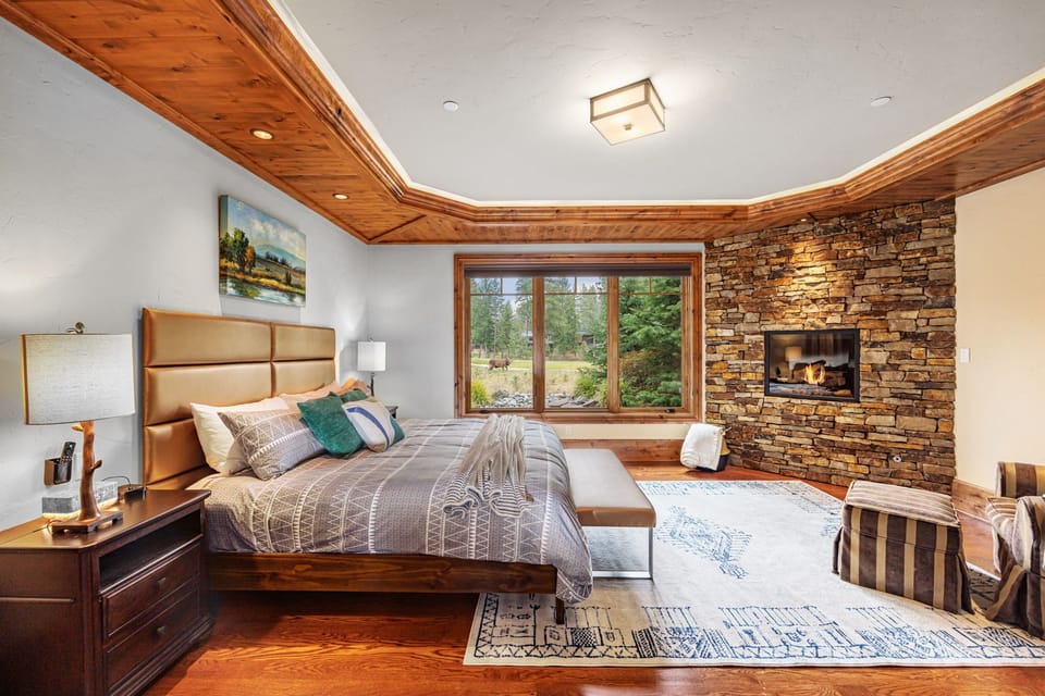 Main Floor - Bedroom 1, Primary Suite King Size Bed + En Suite Bathroom with stacked-stone gas fireplace, wood ceilings, and garden views.