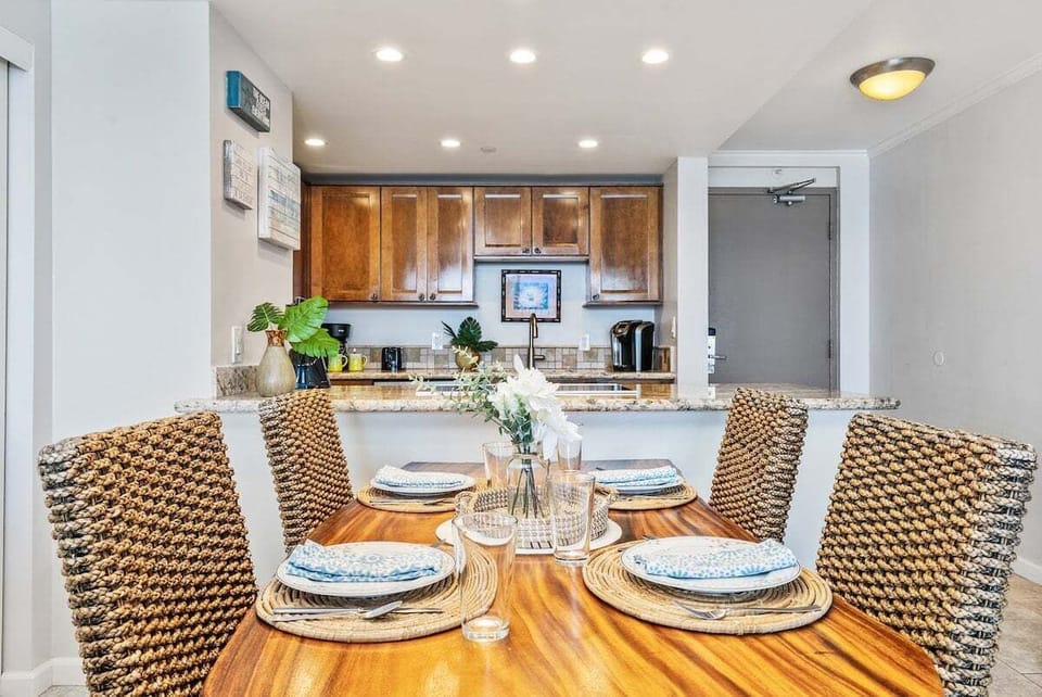 Dining table set for four with woven chairs — gather for laughs, clink glasses, and let the kitchen glow in the background.