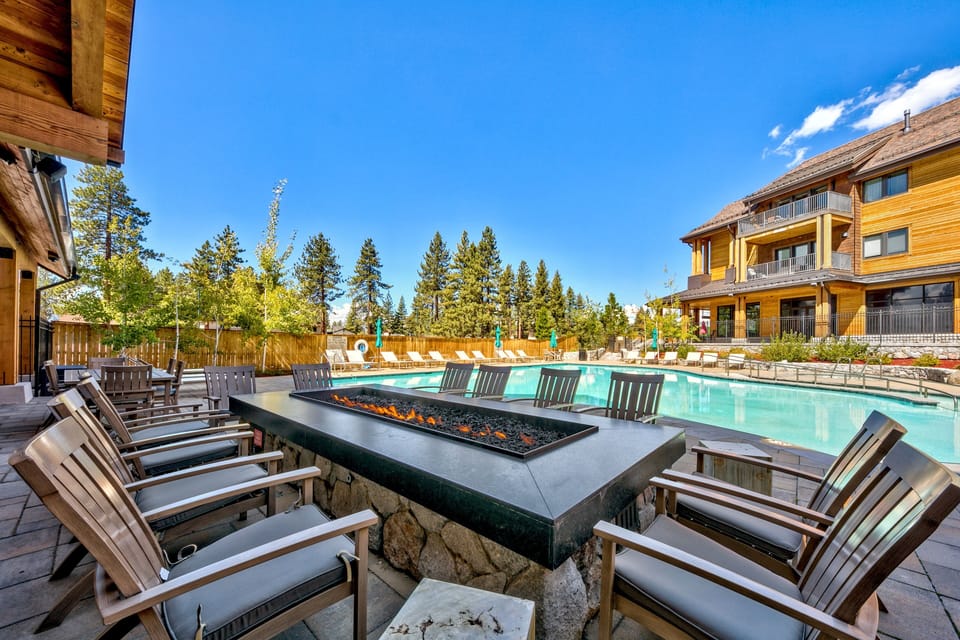 A sparkling pool surrounded by modern alpine lodges and towering evergreens under brilliant blue skies.