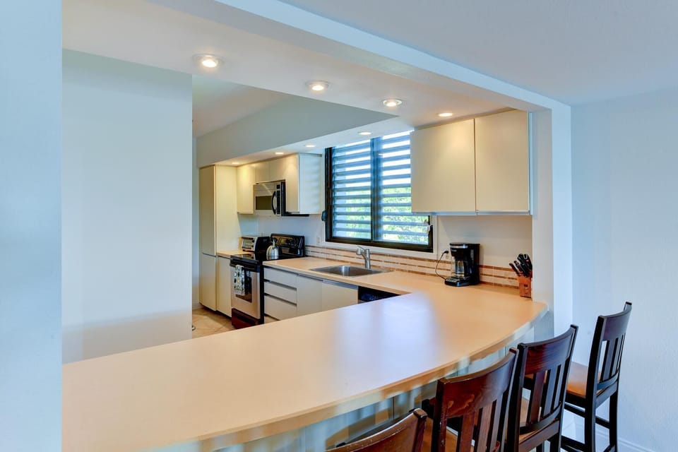 Cream-colored kitchen countertops curve around a breakfast bar with four dark wooden chairs beneath recessed ceiling lights.