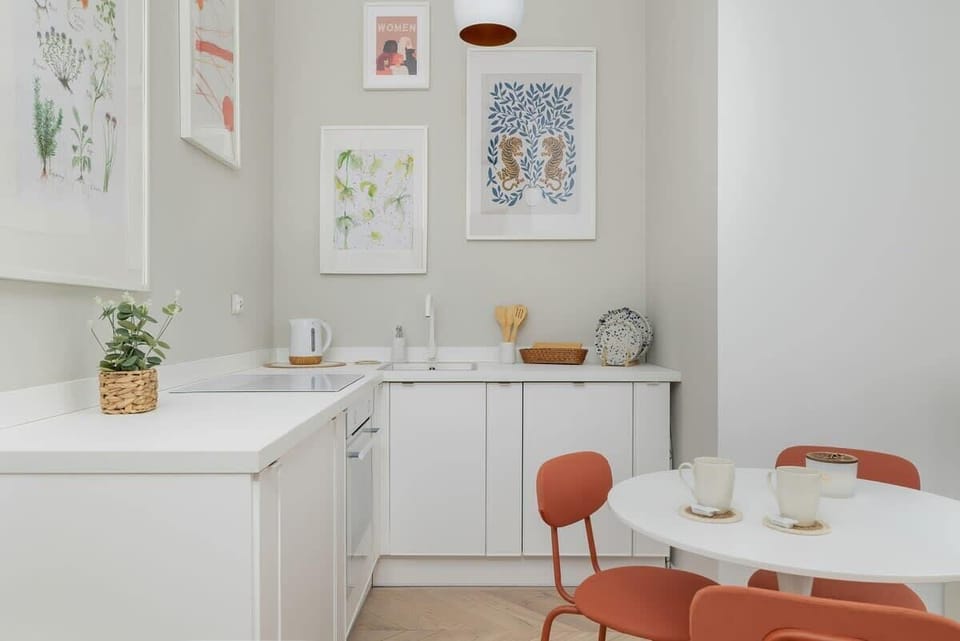 A bright dining zone with a white round table, four orange chairs, and colorful framed prints on the walls.
