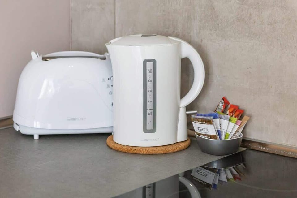 A close-up of a kitchen counter with a white toaster, electric kettle, and a small bowl of tea and coffee.

