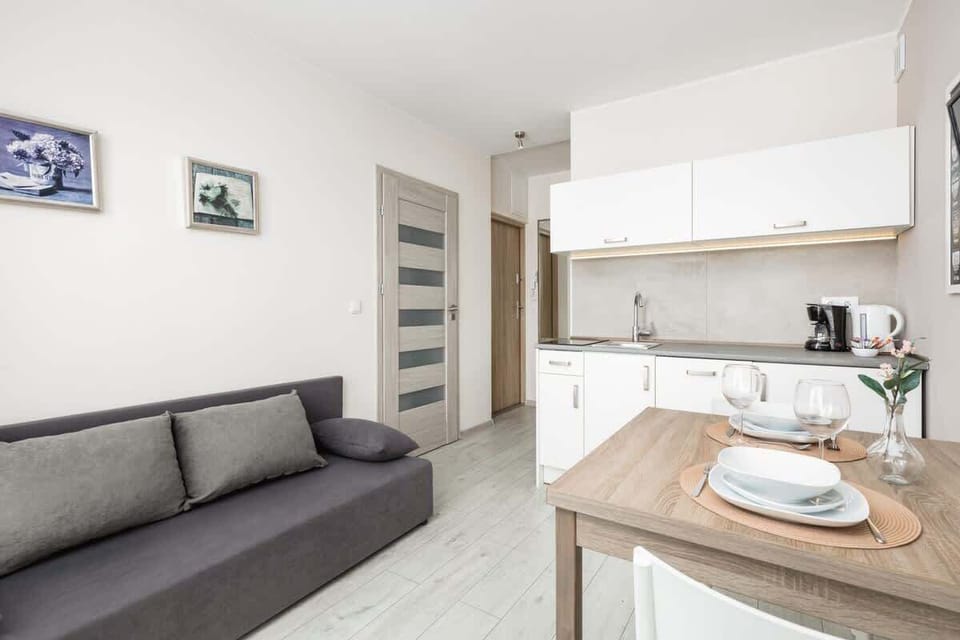 A modern living area with a gray sofa, white kitchen, and a small dining table.


