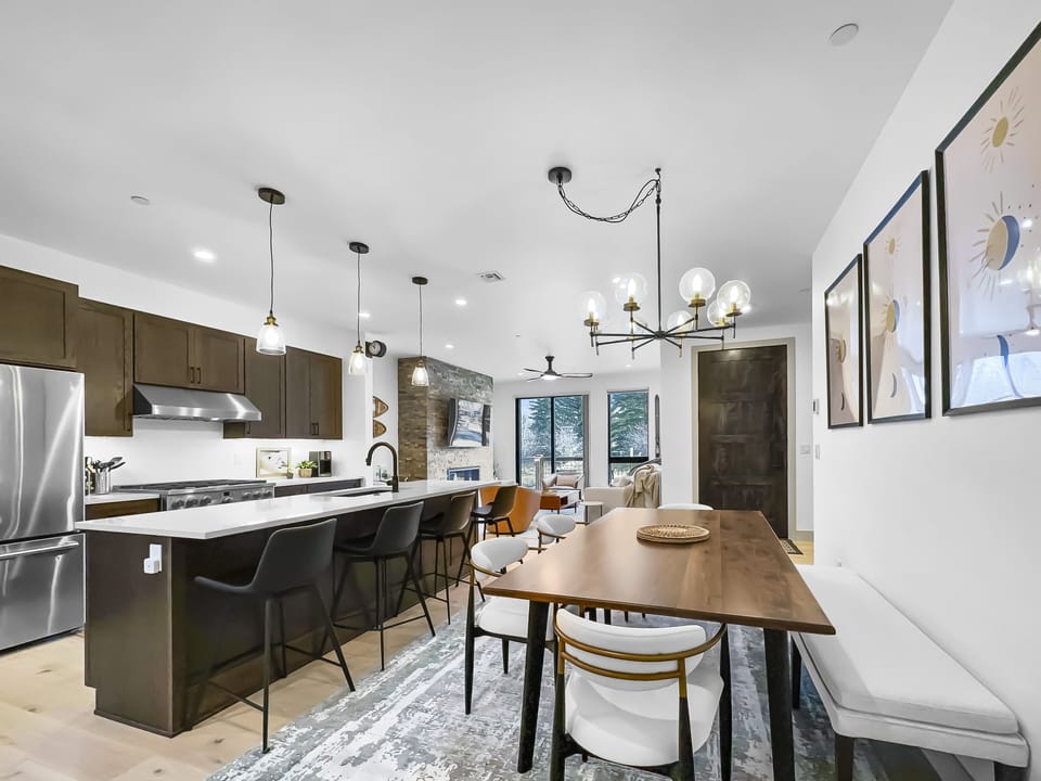 Modern open-concept kitchen and dining area featuring dark cabinetry, stainless steel appliances, an island with barstools, a wooden dining table, and contemporary light fixtures.