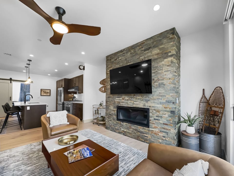 A modern living room with a stone accent wall featuring a mounted TV and fireplace. There are brown leather chairs, a wooden coffee table, and a kitchen with an island visible in the background.