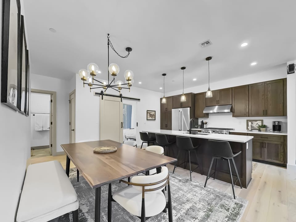A modern kitchen with dark wood cabinets, white countertops, and an island with bar stools, adjacent to a dining area with a wooden table, white and black chairs, and a green patterned rug.