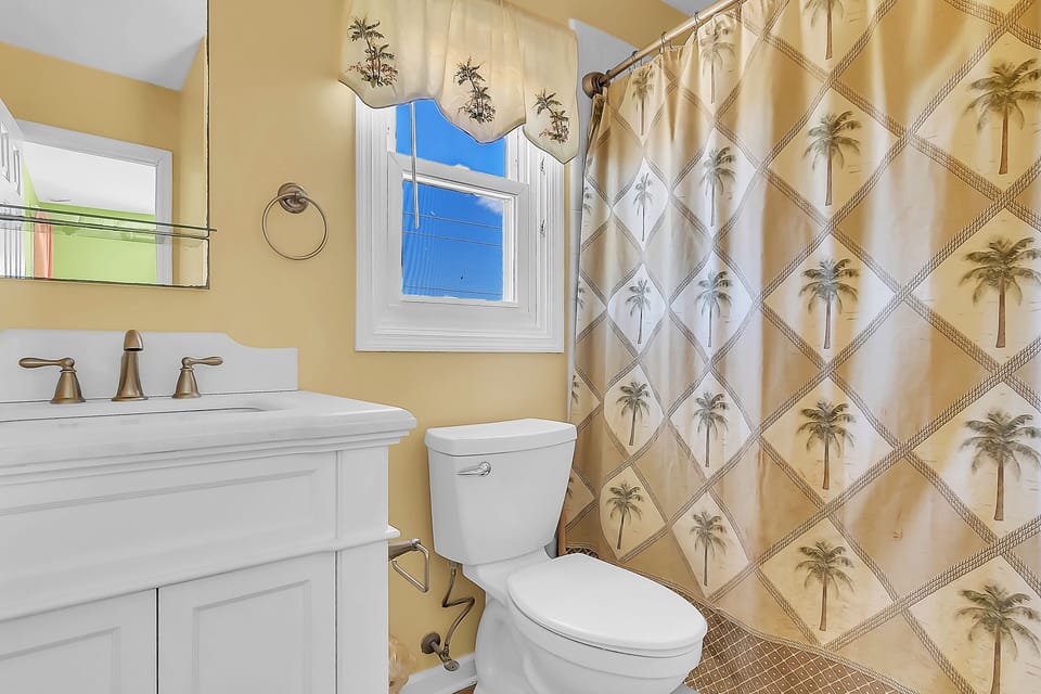 Your tropical-themed bathroom features warm golden tones, a palm-print shower curtain, and bright natural light.