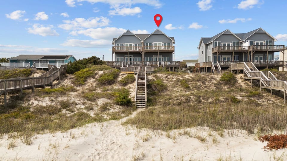 Oceanfront duplex rental with private beach access via wooden staircase, surrounded by coastal dunes and neighboring beach houses.