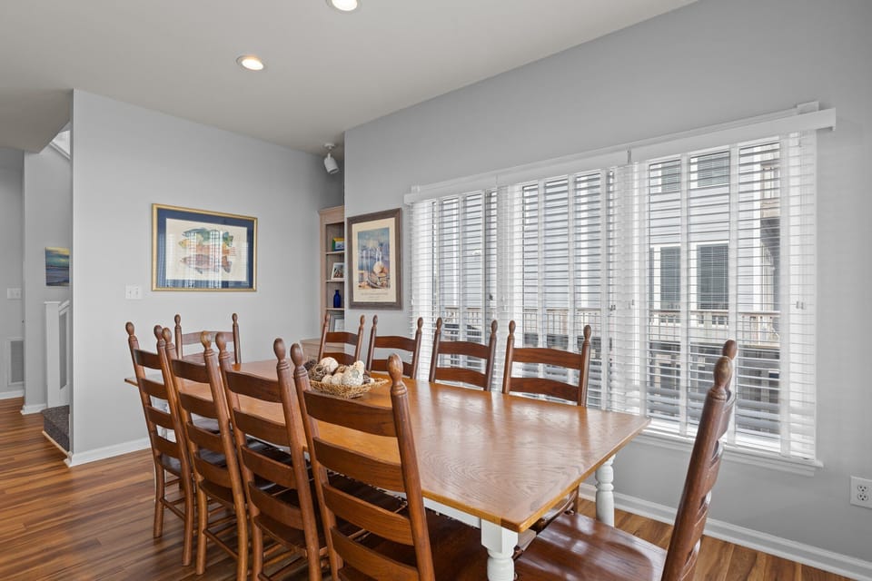Gather around this spacious dining table with warm wood tones and natural light streaming through blinds for memorable meals together.