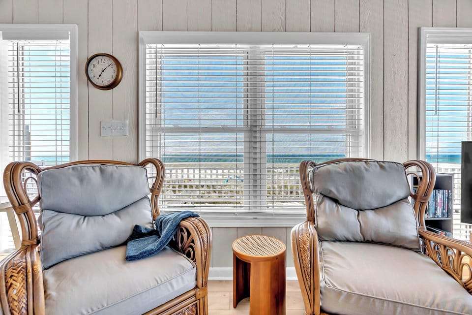 Settle into your coastal-inspired seating area and watch the waves roll in through oversized windows framed by breezy white blinds. Gentle ocean blues meet warm rattan, creating your perfect beachside retreat.