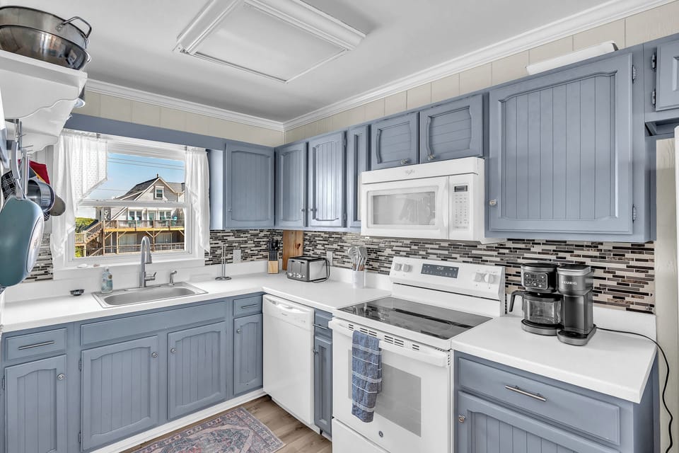 Your fully equipped kitchen features coastal blue cabinetry, crisp white countertops, and modern appliances—perfect for preparing meals while enjoying neighborhood views through bright windows.
