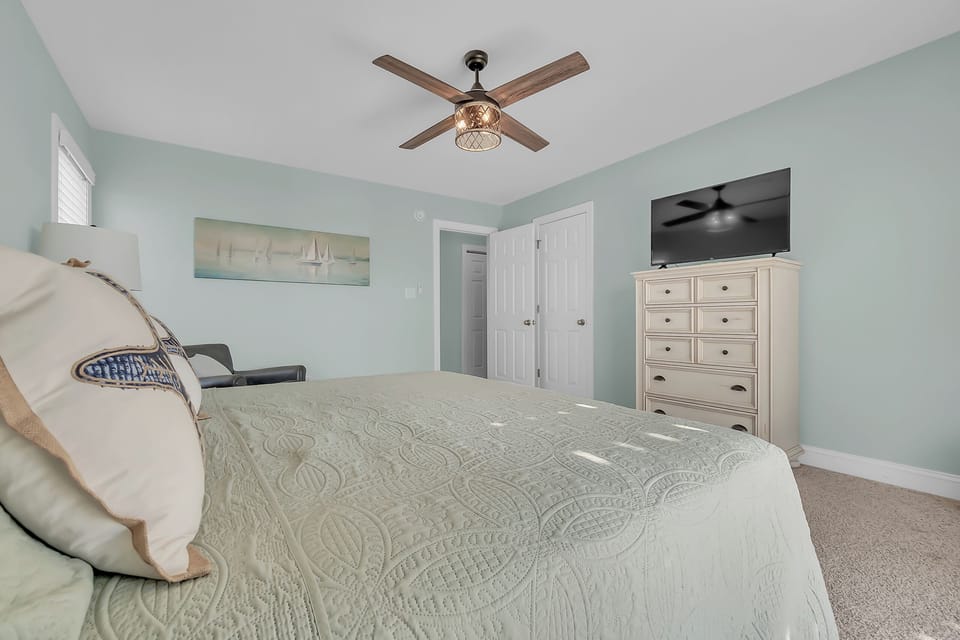 Unwind in your serene bedroom featuring soft mint walls, coastal artwork, and a gentle ceiling fan for perfect comfort.