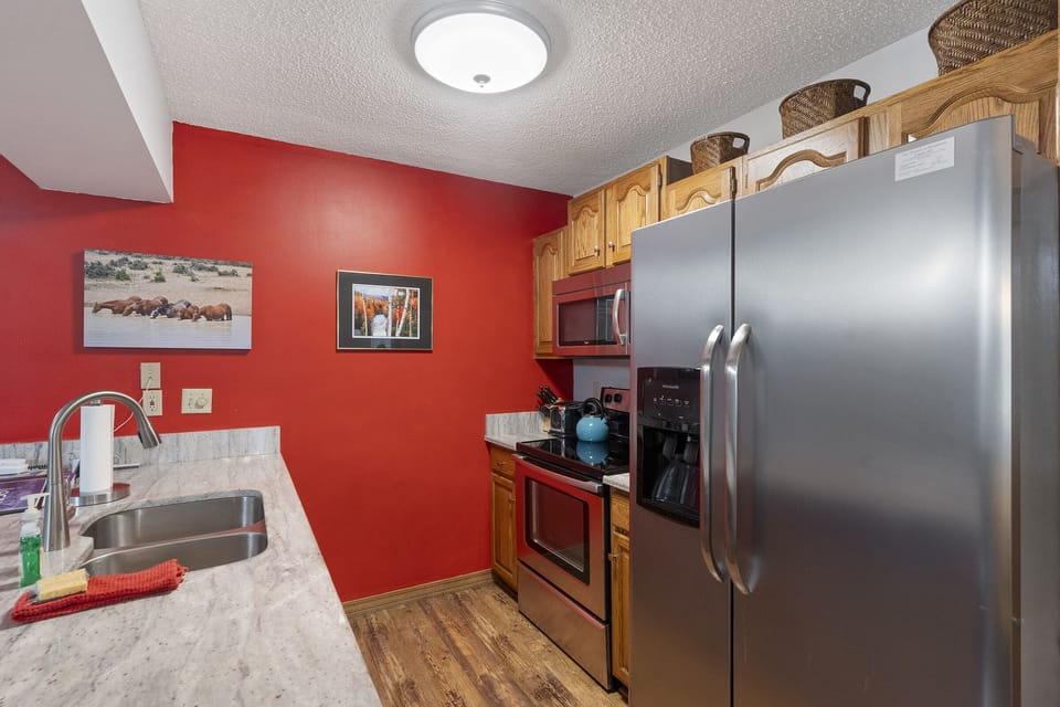 A small kitchen with red walls, stainless steel refrigerator, oven, microwave, and wooden cabinets. Two framed photos hang on the wall above the countertop with a sink and various kitchen items.