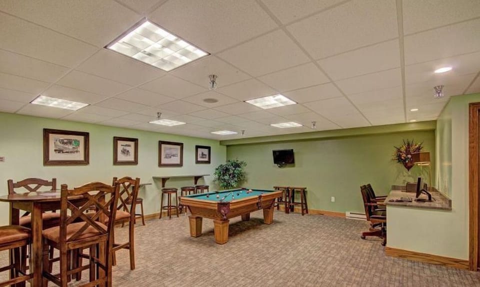 A basement recreation room with a pool table, bar-height tables and chairs, a flat-screen TV, framed artwork, and a bouquet of artificial flowers on a counter. The floor is carpeted and the ceiling has recessed lighting.