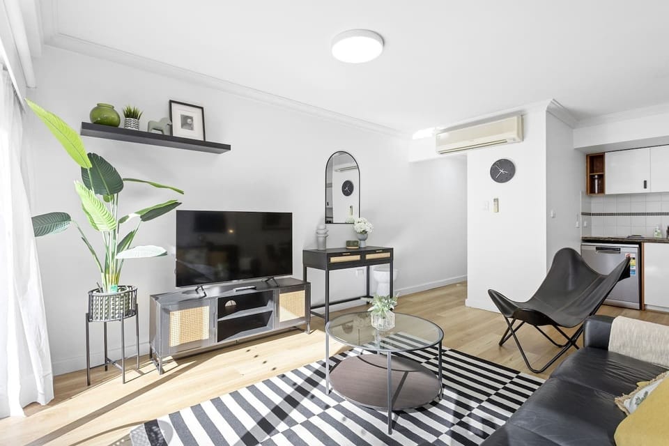 Bright and beautifully furnished living room with a comfortable black leather sofa, stylish decor, and large glass doors opening to a green view. Perfect for relaxation.