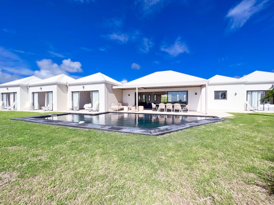 The image depicts a white luxury villa with an outdoor pool, surrounded by loungers and a well-kept garden under a clear blue sky.