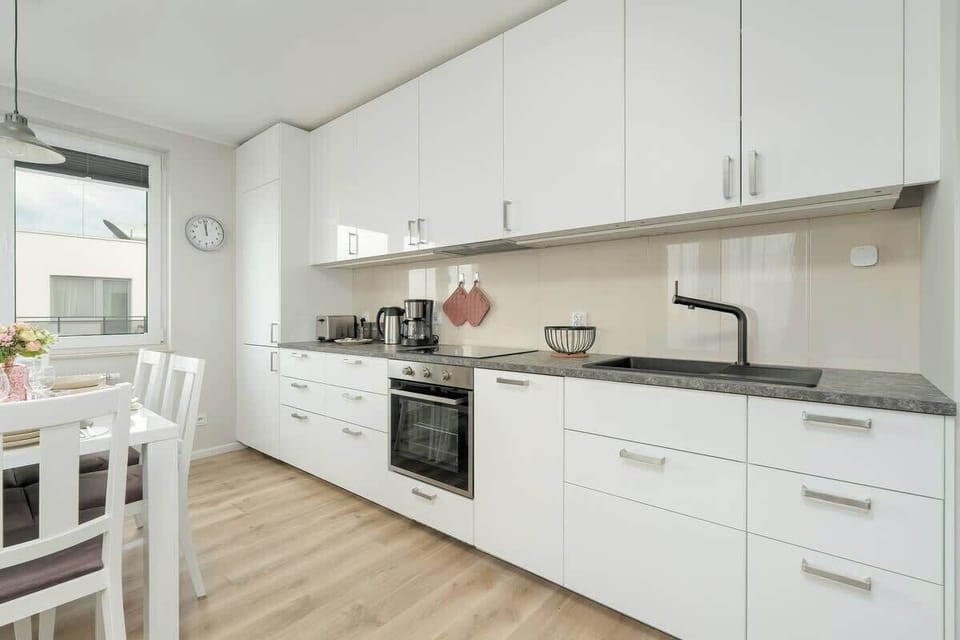 A modern kitchen with a white color scheme. There’s a large table with white chairs, and the kitchen is equipped with sleek appliances and cabinetry. The room is spacious and bright.
