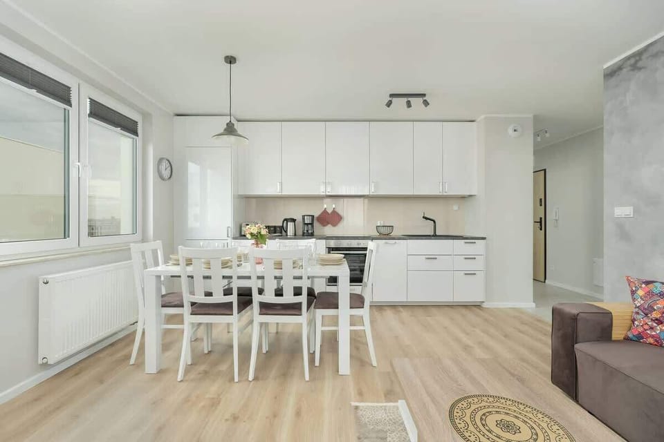 A cozy living and dining area with a light wood floor. There’s a comfortable sofa in the living space and a dining table with white chairs. Large windows bring in natural light.