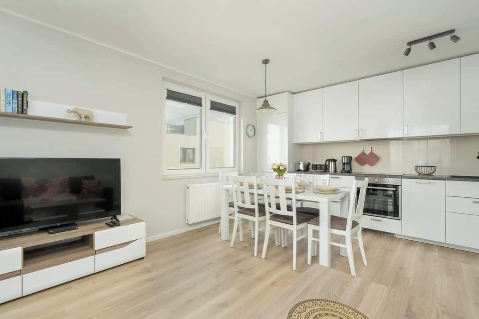 A modern kitchen with a white color scheme. There’s a large table with white chairs, and the kitchen is equipped with sleek appliances and cabinetry. The room is spacious and bright.