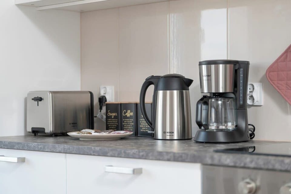 A modern kitchen counter with various small appliances like a kettle and coffee maker. The countertops are clean and minimalistic, reflecting a modern, functional kitchen.