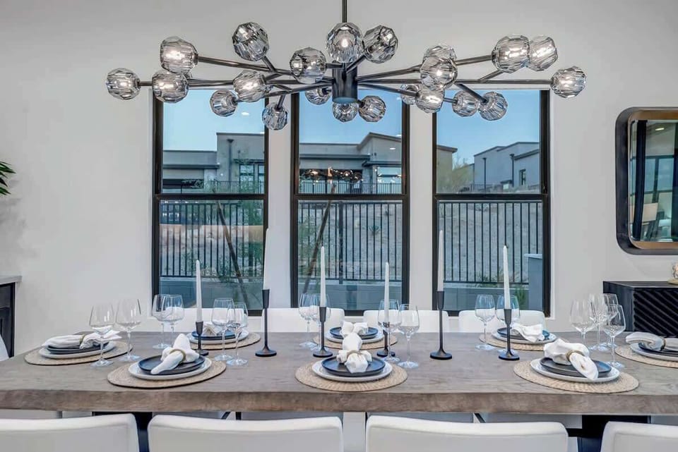 This dining room exudes sophistication with its long table set for six and an elaborate chandelier that adds a touch of glamour. The large windows with black frames let in plenty of natural light, creating a perfect ambiance for formal dinners or special occasions.