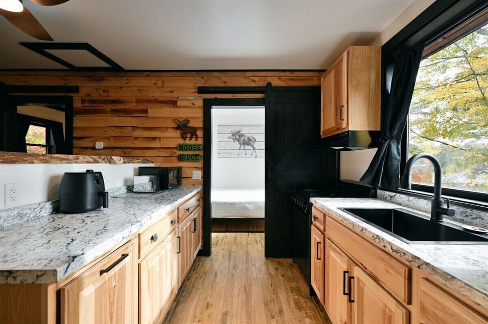 Modern kitchen area with light wood cabinets, marbled countertops, a black sink, and a view into a bedroom with a "Moose Ridge" sign on the wall.