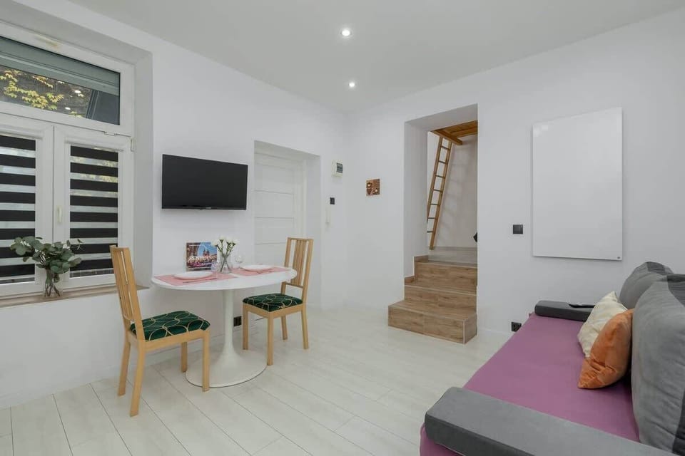 Living room area with a sofa, white walls, and wooden elements. There are stairs leading up to an additional space, and the overall ambiance is modern and clean.