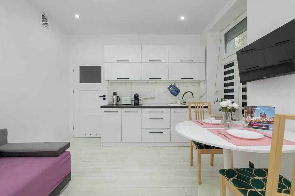  Kitchen area with a white color palette. It features modern cabinetry, a white dining table, and a pink sofa to the side.