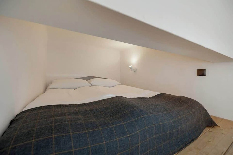Bedroom with a bed tucked under a low ceiling or lofted area, giving a cozy feel to the space. The bed has a dark comforter, and the walls are white.