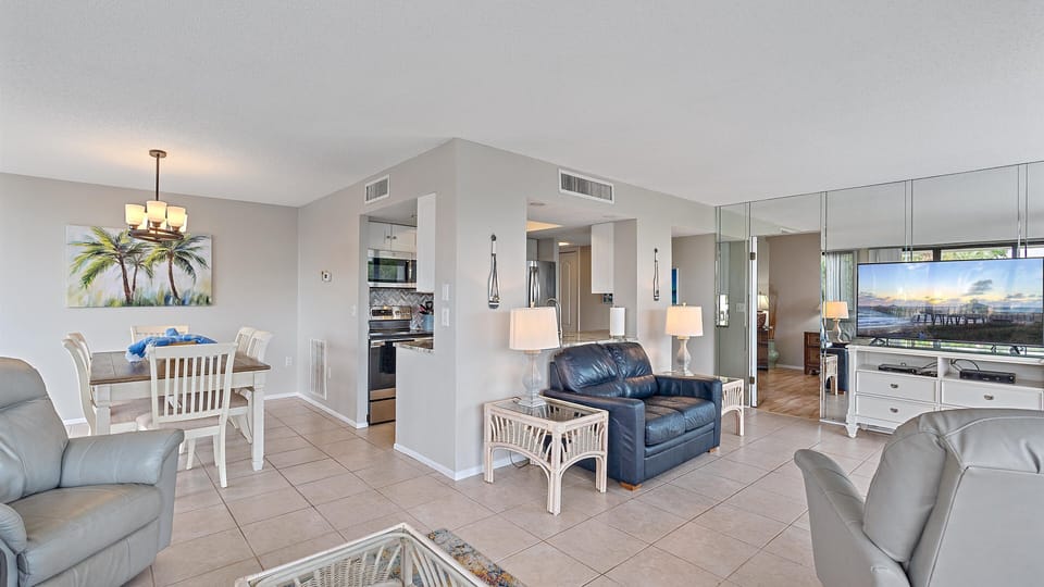 Modern open-plan living room and dining area with tiled floors, light-colored walls, leather armchairs, a dining table, and a kitchen in the background. A large mirror and television are on the right wall.