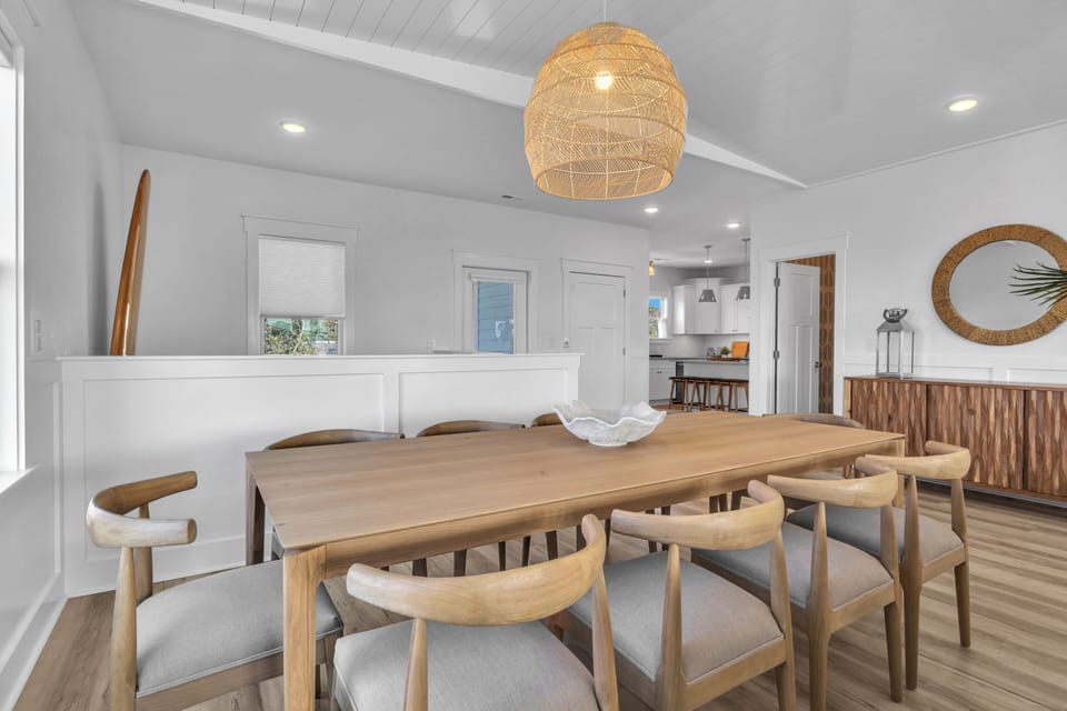 Bright dining area with wood table, 6 chairs, woven pendant light, plank floors, windows, stair rail, deck door, surfboard decor, plant, and console table with stools.