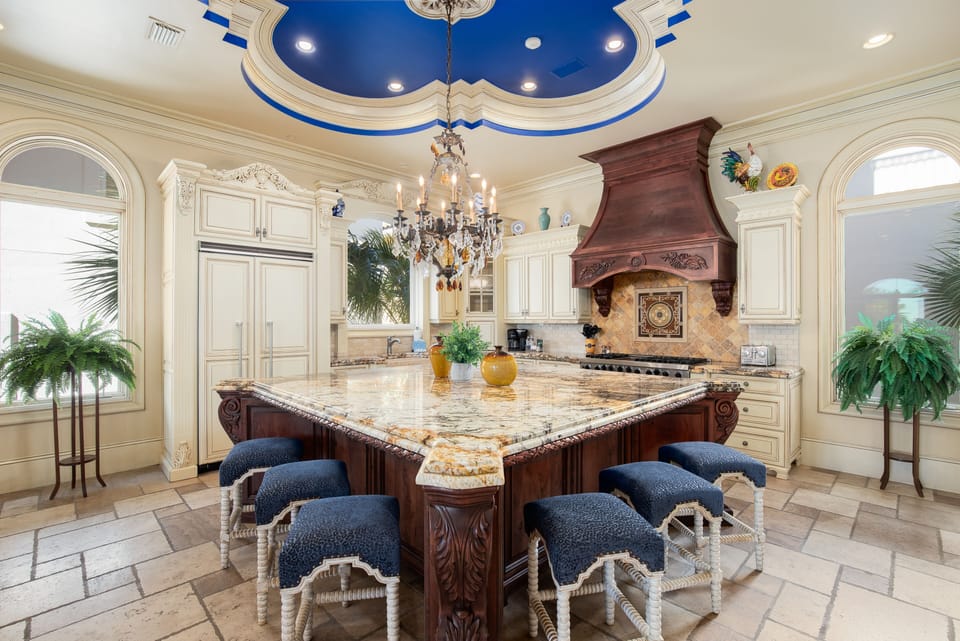 Step into your grand culinary sanctuary where marble countertops and crystal chandeliers create an elegant cooking experience beneath stunning blue ceiling details.