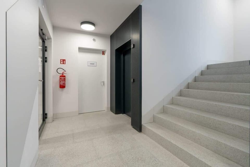 A clean and modern building staircase with neutral tiles, dark grey doors, and a minimalist design, accompanied by a fire extinguisher for safety.
