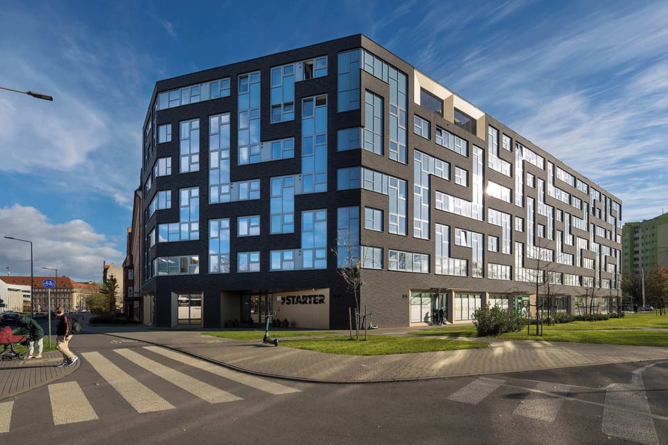 A view of a modern apartment building with an intricate, geometric design. The building features large windows, a clean facade, and well-maintained greenery surrounding it.