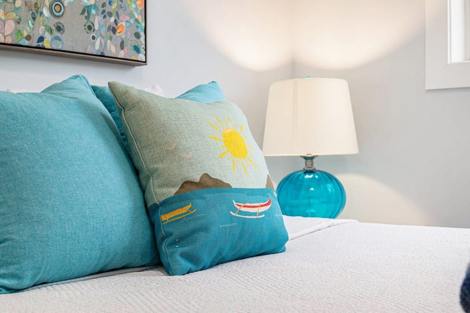 Turquoise pillows and a glass bedside lamp beside white bedding and a colorful cushion.