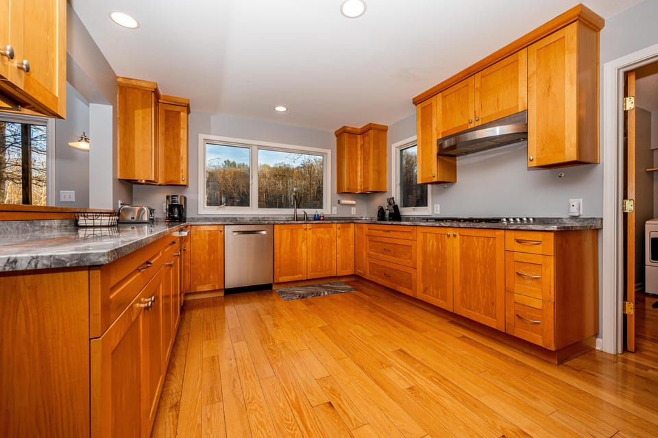 Spacious kitchen with honey-toned wood cabinets, gray marble countertops, stainless dishwasher, and wood flooring.