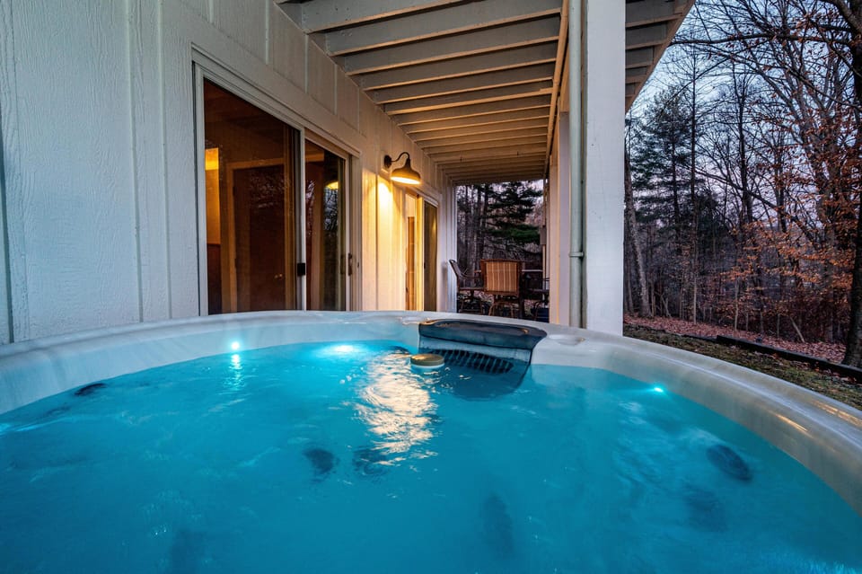 Blue-lit hot tub sits under a covered porch with white vertical siding and overlooks a wooded yard at dusk.