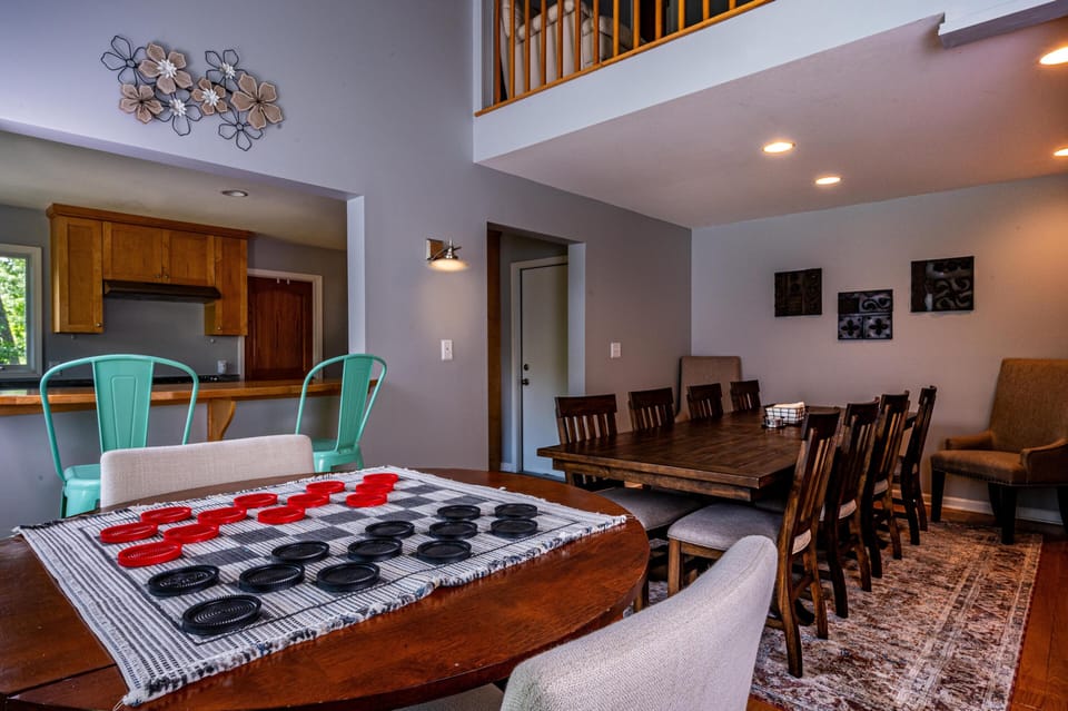 Game-ready round wood table with checkerboard pieces, teal metal stools, and a long dining table on patterned rug.